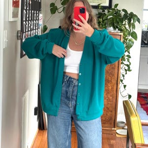 Può includere: Cardigan verde acqua con chiusura a bottoni. È abbinato a jeans in denim chiaro. L'outfit è ripreso in uno specchio, con uno scorcio di un mobile in legno e un'opera d'arte incorniciata sullo sfondo.