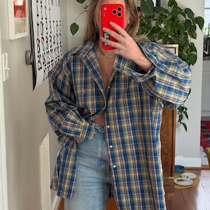 May include: A button-down plaid shirt in blue, yellow, and red. The shirt has long sleeves and a classic collar. The person is wearing light blue jeans. The shirt is unbuttoned at the waist.
