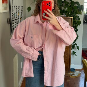 Puede incluir: Una camisa rosa claro con botones y un pequeño logotipo bordado en el bolsillo del pecho. La camisa se combina con unos vaqueros azules. El conjunto es informal y cómodo, adecuado para el uso diario.