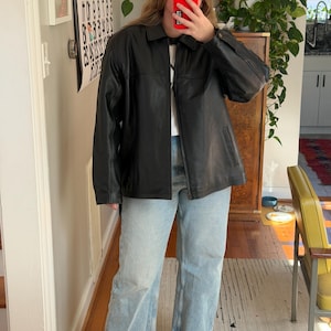 May include: A black leather jacket with a collared neckline and a zipper closure. The jacket has a classic design with visible stitching details. The person is wearing light blue jeans and black platform boots.