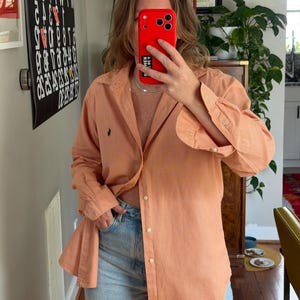 May include: A peach-colored button-down shirt with a small, dark logo on the chest. The shirt has a subtle checkered pattern and long sleeves. The person is wearing light blue jeans.