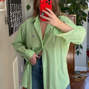May include: A light green button-down shirt with a small blue logo on the chest. The shirt is paired with blue jeans. The shirt has long sleeves and a classic collar. The fabric appears to have a subtle striped pattern.