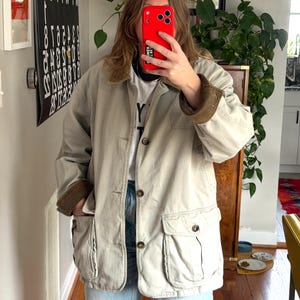 May include: A tan jacket with four front pockets, button closures, and a corduroy collar and cuffs. The jacket is worn over a white t-shirt and blue jeans. The jacket is a neutral color and appears to be made of a durable material.