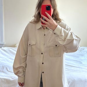 Puede incluir: Una camisa de botones de manga larga de color crema con dos bolsillos grandes con solapa en la parte delantera. La camisa tiene un corte holgado y está hecha de una tela ligera.
