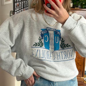 May include: A light gray sweatshirt with long sleeves. The front features a blue and white emblem with the text "UNC ATHLETIC CLUB NORTH CAROLINA" in a collegiate style. The sweatshirt is paired with blue jeans.