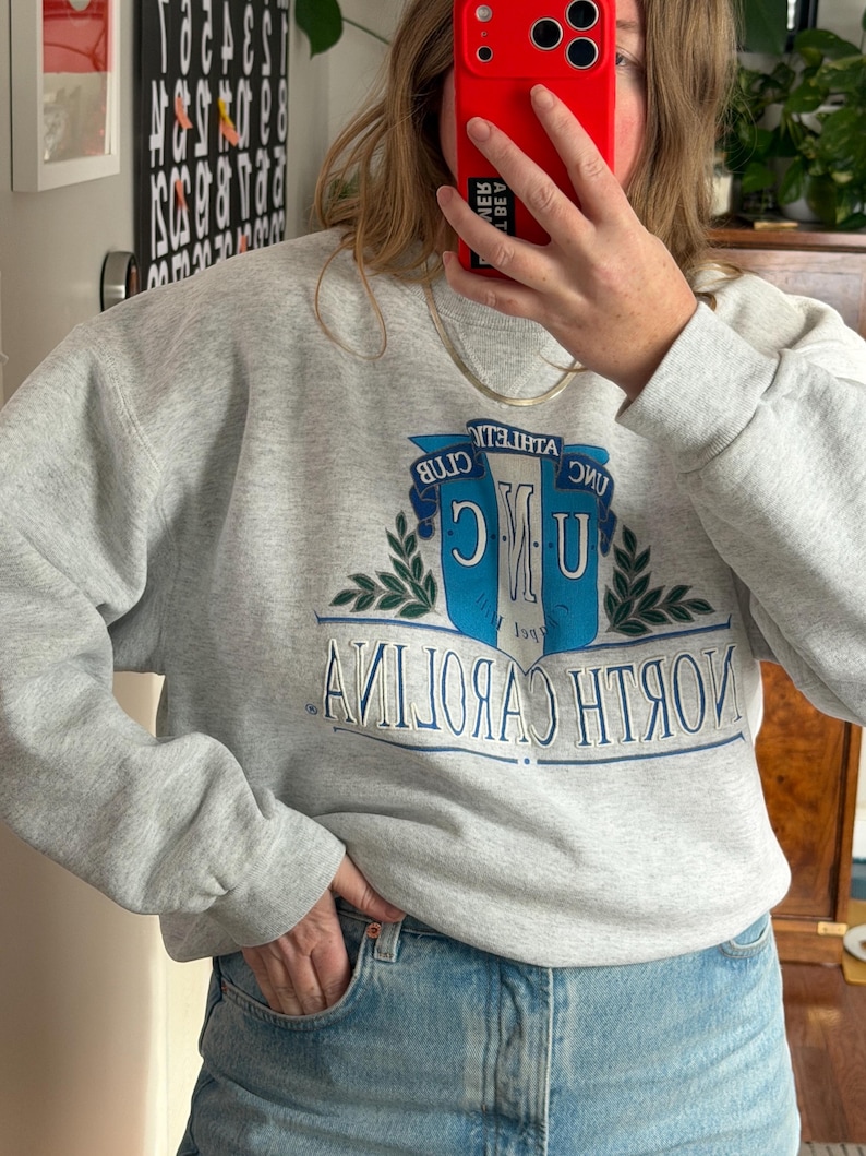 May include: A light gray sweatshirt with long sleeves. The front features a blue and white emblem with the text "UNC ATHLETIC CLUB NORTH CAROLINA" in a collegiate style. The sweatshirt is paired with blue jeans.