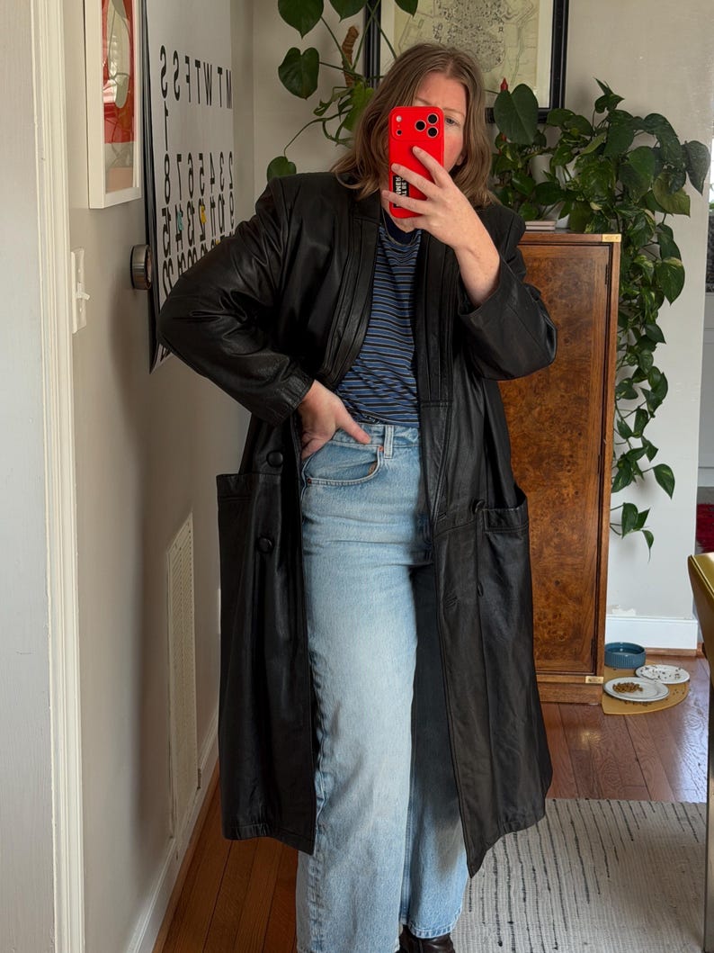 May include: A long black leather coat with a button closure and two front pockets. The coat is worn over a blue and white striped shirt and light blue jeans. The coat appears to be knee-length.
