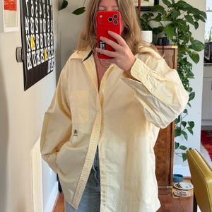 May include: A long-sleeved, pale yellow button-down shirt featuring a classic collar, a single chest pocket, and a small embroidered logo on the lower right side. The shirt is worn untucked over light blue denim jeans with a relaxed fit.