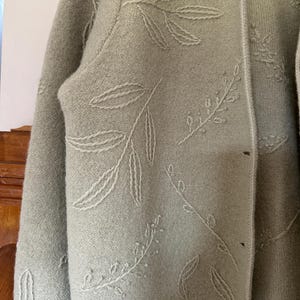 May include: Sage green cardigan with a leaf pattern embroidered in a lighter shade. The cardigan has a collared neckline and button closure. The fabric appears to be a soft knit, suitable for layering.