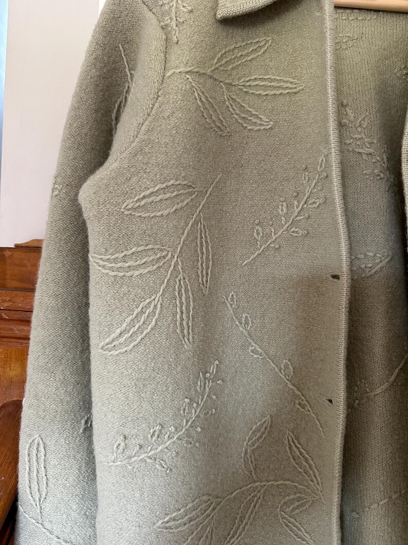May include: Sage green cardigan with a leaf pattern embroidered in a lighter shade. The cardigan has a collared neckline and button closure. The fabric appears to be a soft knit, suitable for layering.