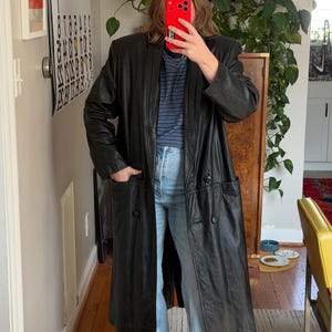 May include: A long, black leather coat with a double-breasted design. The coat features large pockets and a classic lapel. The person is wearing blue jeans and a striped shirt. The coat appears to be vintage.