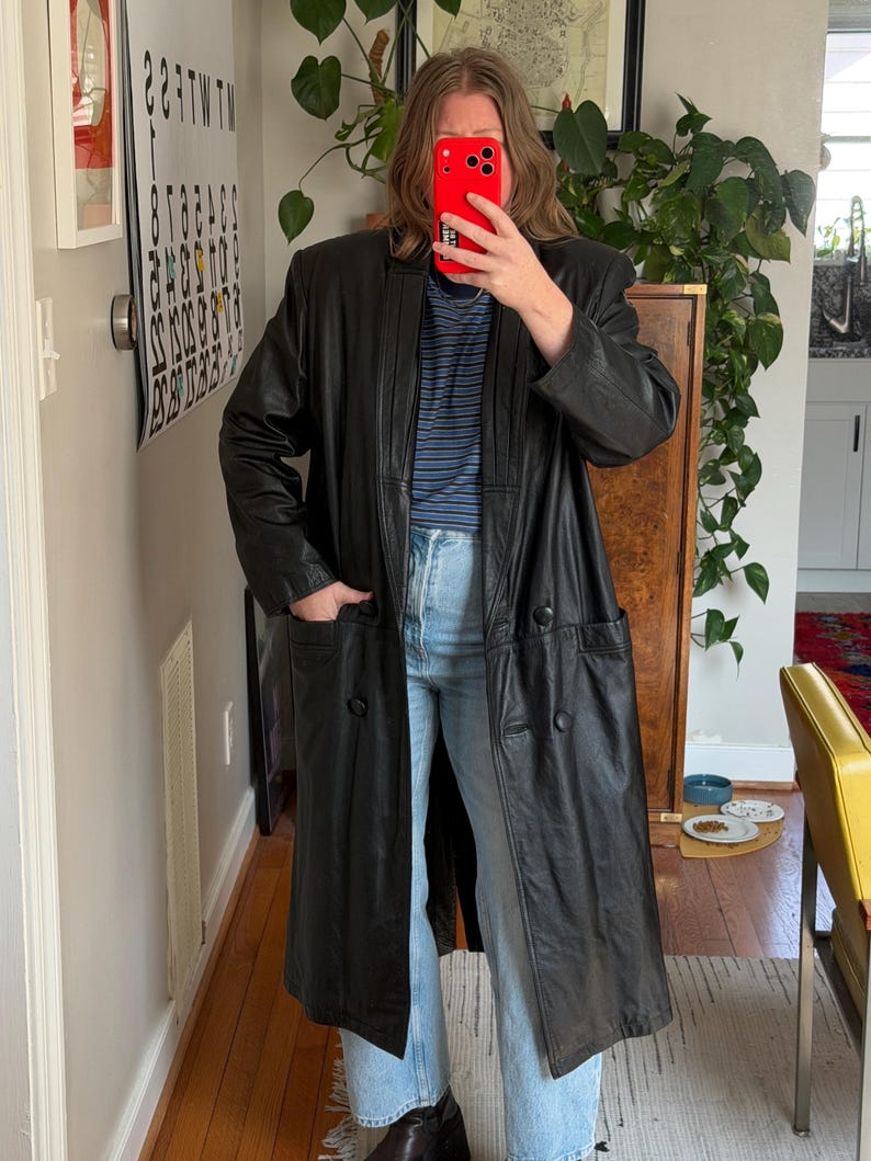 May include: A long, black leather coat with a double-breasted design. The coat features large pockets and a classic lapel. The person is wearing blue jeans and a striped shirt. The coat appears to be vintage.