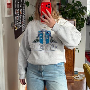May include: A light gray sweatshirt with the text "NORTH CAROLINA" in blue and white. The sweatshirt has a crew neck and long sleeves. The person is wearing light blue jeans.