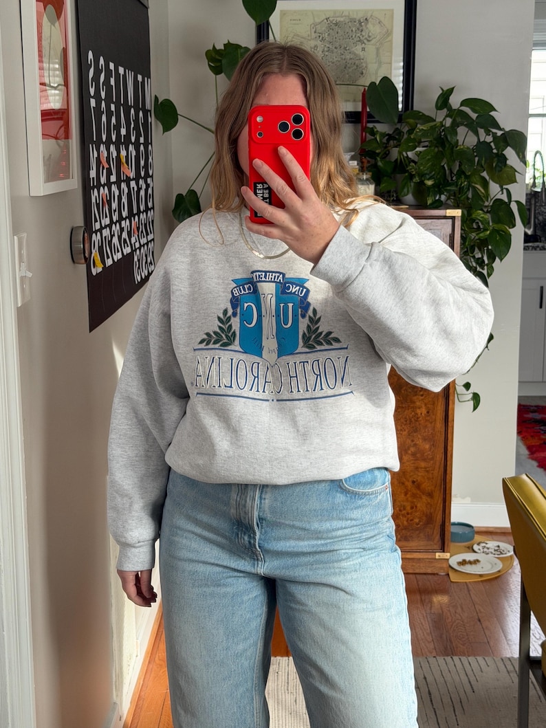 May include: A light gray sweatshirt with the text "NORTH CAROLINA" in blue and white. The sweatshirt has a crew neck and long sleeves. The person is wearing light blue jeans.