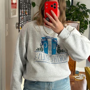 May include: A light gray sweatshirt with the text "NORTH CAROLINA" in blue and white, and the letters "UNC" in a stylized design. The sweatshirt has long sleeves and a crew neck.