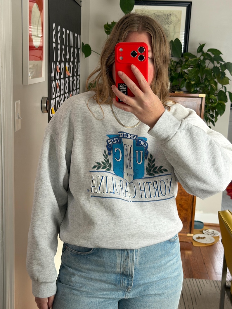 May include: A light gray sweatshirt with the text "NORTH CAROLINA" in blue and white, and the letters "UNC" in a stylized design. The sweatshirt has long sleeves and a crew neck.