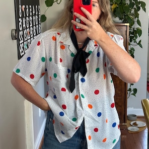 May include: White button-down shirt with a polka dot pattern in red, blue, green, and orange. The shirt has short sleeves and a black tie. The model is wearing blue jeans.