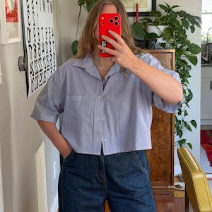 May include: A cropped, short-sleeved button-down shirt with vertical blue and white stripes. The shirt has a single chest pocket and is paired with dark wash denim jeans. The model is holding a red phone.
