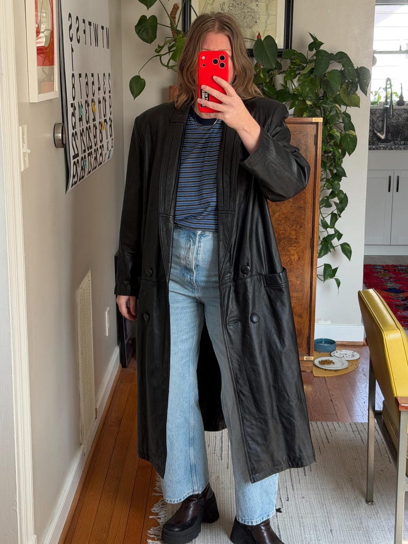 May include: A long, black leather coat with a double-breasted design. The coat is worn over a blue and white striped shirt and light wash jeans. The person is wearing platform boots.