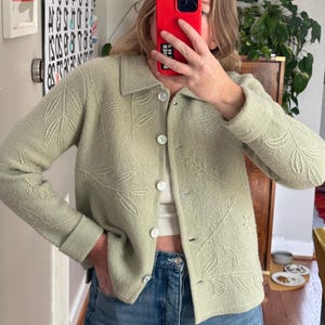 May include: A light green button-up cardigan with embroidered leaf designs. The cardigan has a collared neckline and is buttoned up. The sleeves are cuffed. The cardigan is paired with blue jeans.