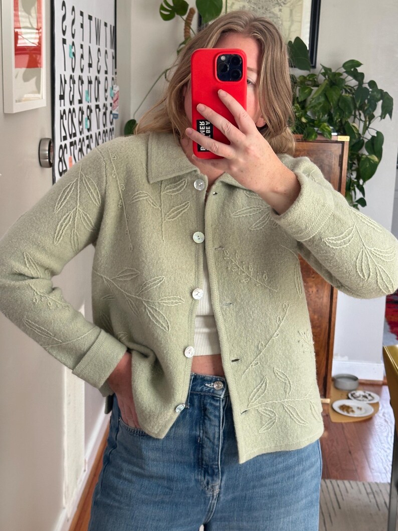 May include: A light green button-up cardigan with embroidered leaf designs. The cardigan has a collared neckline and is buttoned up. The sleeves are cuffed. The cardigan is paired with blue jeans.
