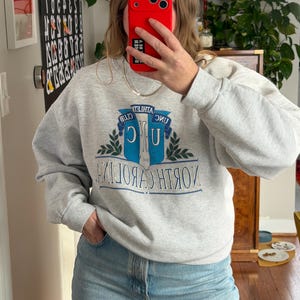 May include: A light gray sweatshirt with the text "UNC" and "North Carolina" in blue and white. The sweatshirt has a crew neck and long sleeves. The image also shows blue jeans.