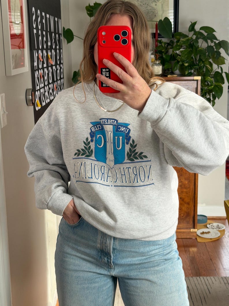 May include: A light gray sweatshirt with the text "UNC" and "North Carolina" in blue and white. The sweatshirt has a crew neck and long sleeves. The image also shows blue jeans.