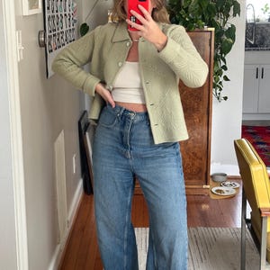 May include: A light green jacket with leaf-like embroidery, paired with a white top and blue jeans. The jacket has a button-up front and a collar. The jeans are wide-legged. The outfit is casual and stylish.