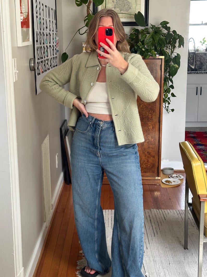 May include: A light green jacket with leaf-like embroidery, paired with a white top and blue jeans. The jacket has a button-up front and a collar. The jeans are wide-legged. The outfit is casual and stylish.