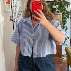 May include: A light blue and white striped cropped shirt with short sleeves and a button-down front. The shirt has a collared neckline and a single pocket. The shirt is paired with dark blue denim jeans.