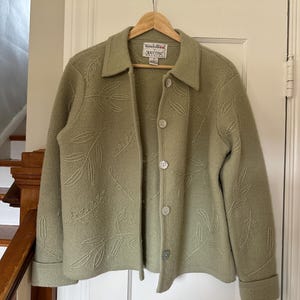 May include: A sage green wool jacket with leaf embroidery. The jacket has a collared neckline, button closure, and long sleeves with folded cuffs. The label reads "Wonderwool Knitting".