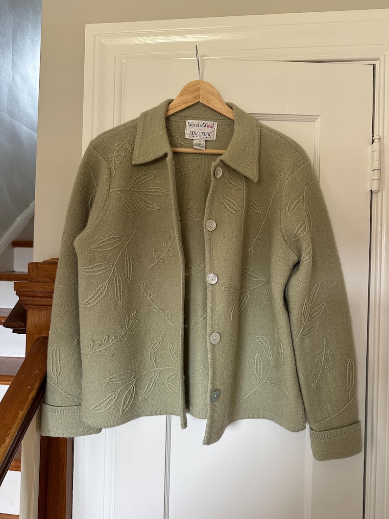 May include: A sage green wool jacket with leaf embroidery. The jacket has a collared neckline, button closure, and long sleeves with folded cuffs. The label reads "Wonderwool Knitting".