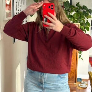 Puede incluir: Un su&eacute;ter burdeos de manga larga con cuello con cremallera. El su&eacute;ter se combina con vaqueros de mezclilla azul claro. La persona sostiene un tel&eacute;fono rojo frente a su rostro.