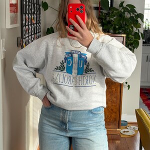 May include: A heather gray sweatshirt with the text "UNC ATHLETIC CLUB NORTH CAROLINA" in blue and white. The sweatshirt has a crew neck and long sleeves. The person in the image is wearing light blue jeans.