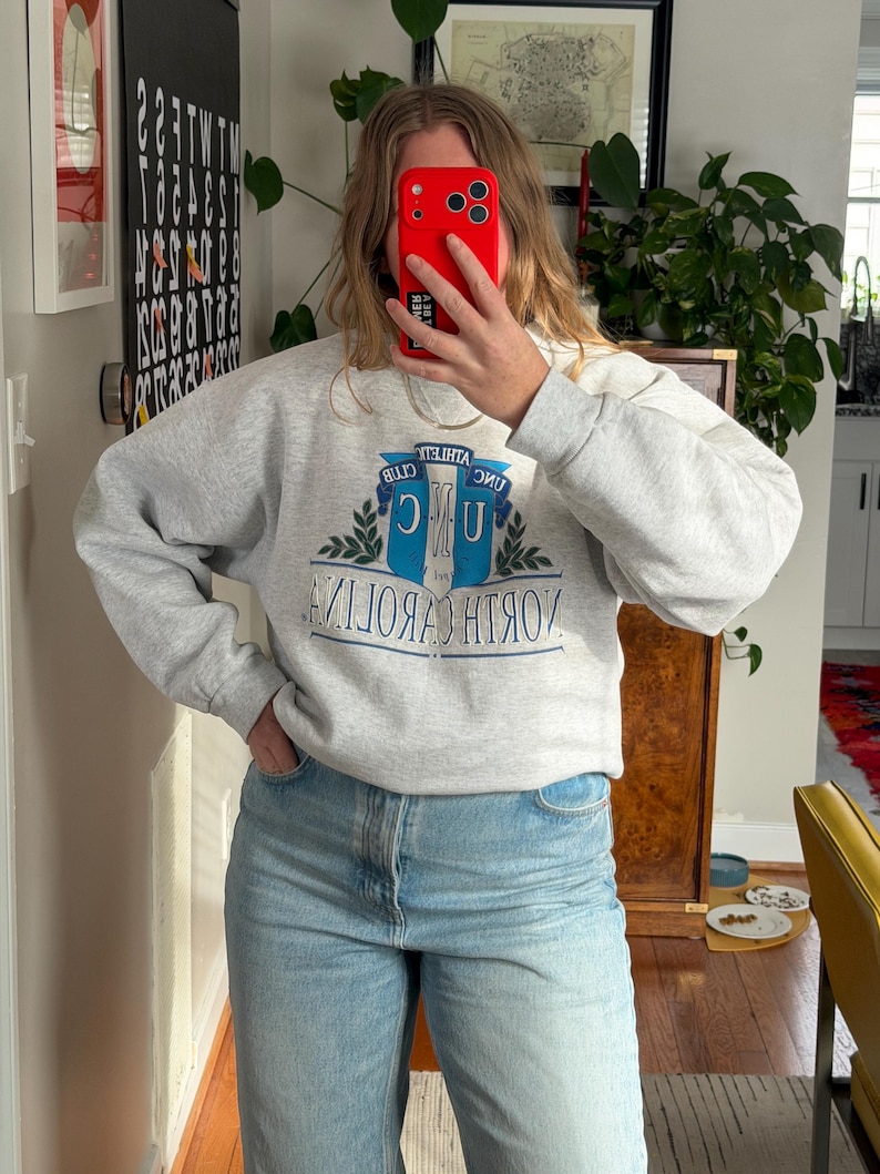 May include: A heather gray sweatshirt with the text "UNC ATHLETIC CLUB NORTH CAROLINA" in blue and white. The sweatshirt has a crew neck and long sleeves. The person in the image is wearing light blue jeans.
