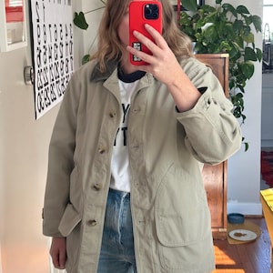 May include: A light tan jacket with a button-up front, two large front pockets, and a dark green collar. The jacket is worn over a white t-shirt and blue jeans. The person is holding a red phone.