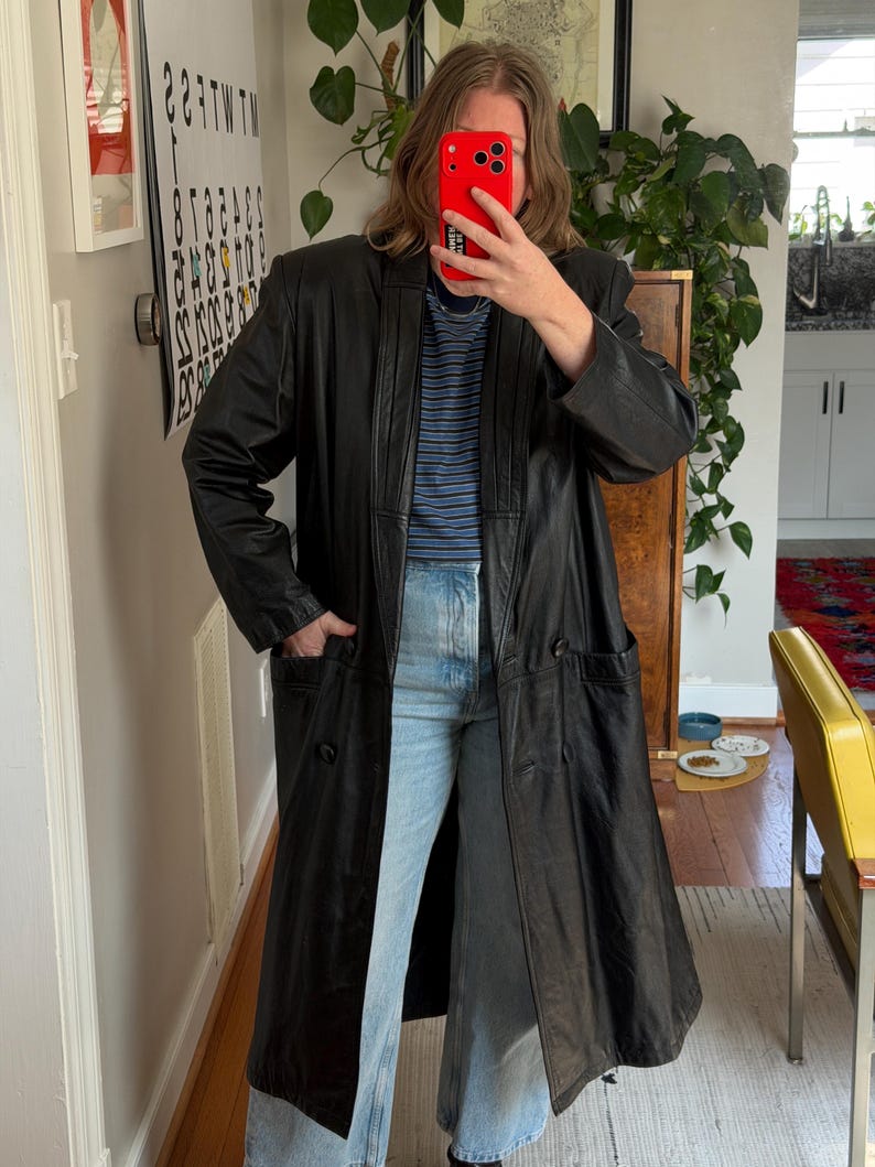 May include: A long, black leather coat with a double-breasted design. The coat features two front pockets and a notched collar. The person in the image is wearing a blue and white striped shirt and light blue jeans.