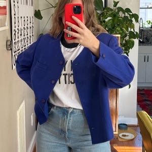 May include: A vibrant blue blazer with a round collar and button fastening. The blazer is worn over a white t-shirt and light blue jeans. The t-shirt has text on it. The person is holding a red phone.