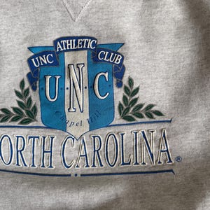 May include: A heather gray sweatshirt featuring a blue and white emblem with the text "UNC Athletic Club" and "North Carolina". The design includes laurel branches and a shield shape.