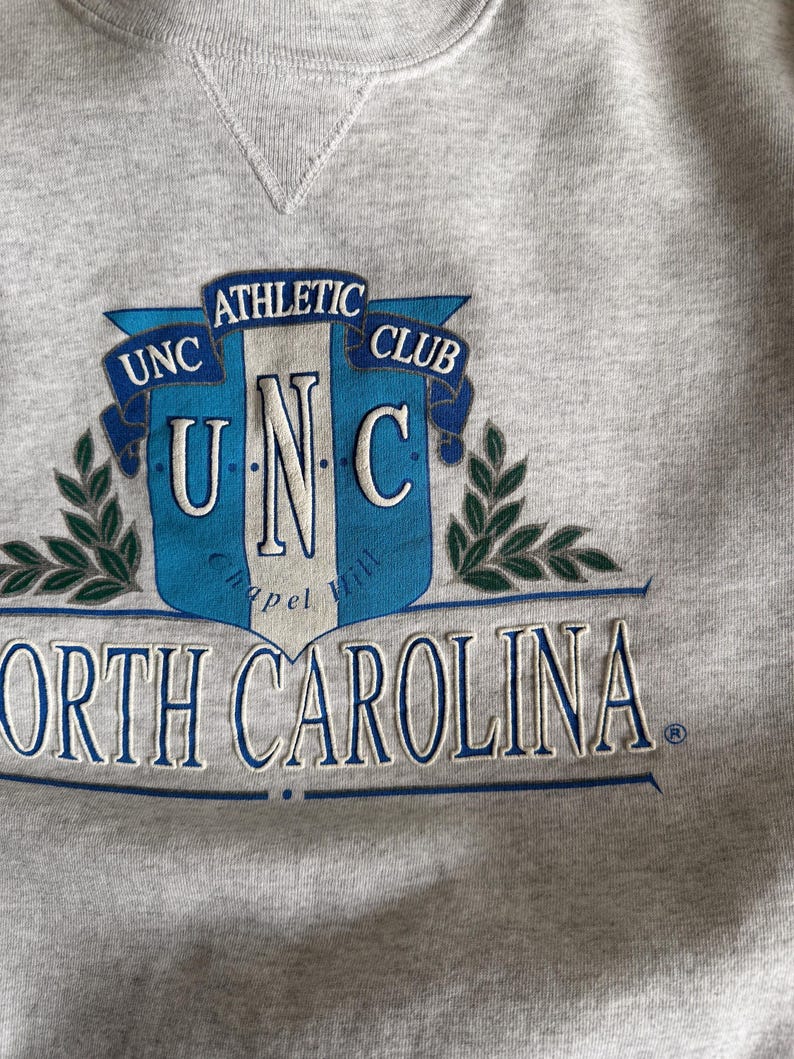 May include: A heather gray sweatshirt featuring a blue and white emblem with the text "UNC Athletic Club" and "North Carolina". The design includes laurel branches and a shield shape.