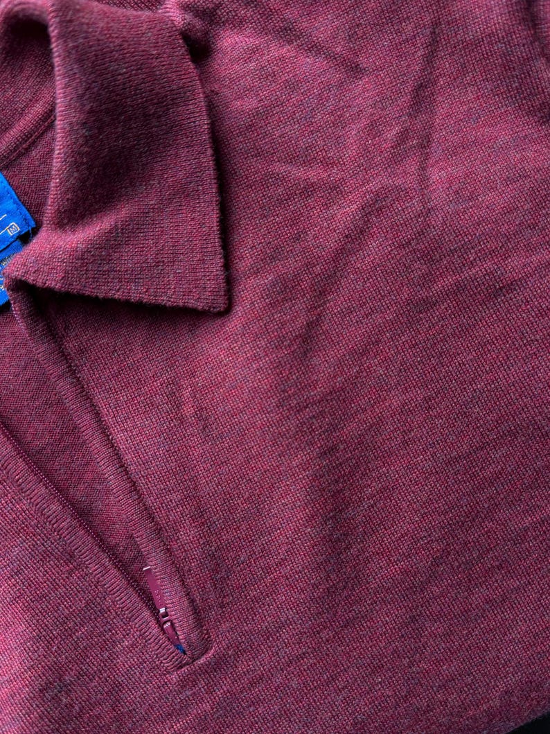 Puede incluir: Primer plano de un su&eacute;ter de punto color burdeos. El su&eacute;ter tiene cuello y cierre de cremallera parcial. La tela tiene un aspecto texturizado.