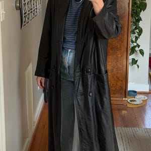 May include: A long, black leather coat with a double-breasted design. The coat has a notched collar, two front pockets, and a long, flowing silhouette. The coat is worn over a blue and white striped shirt and blue jeans.