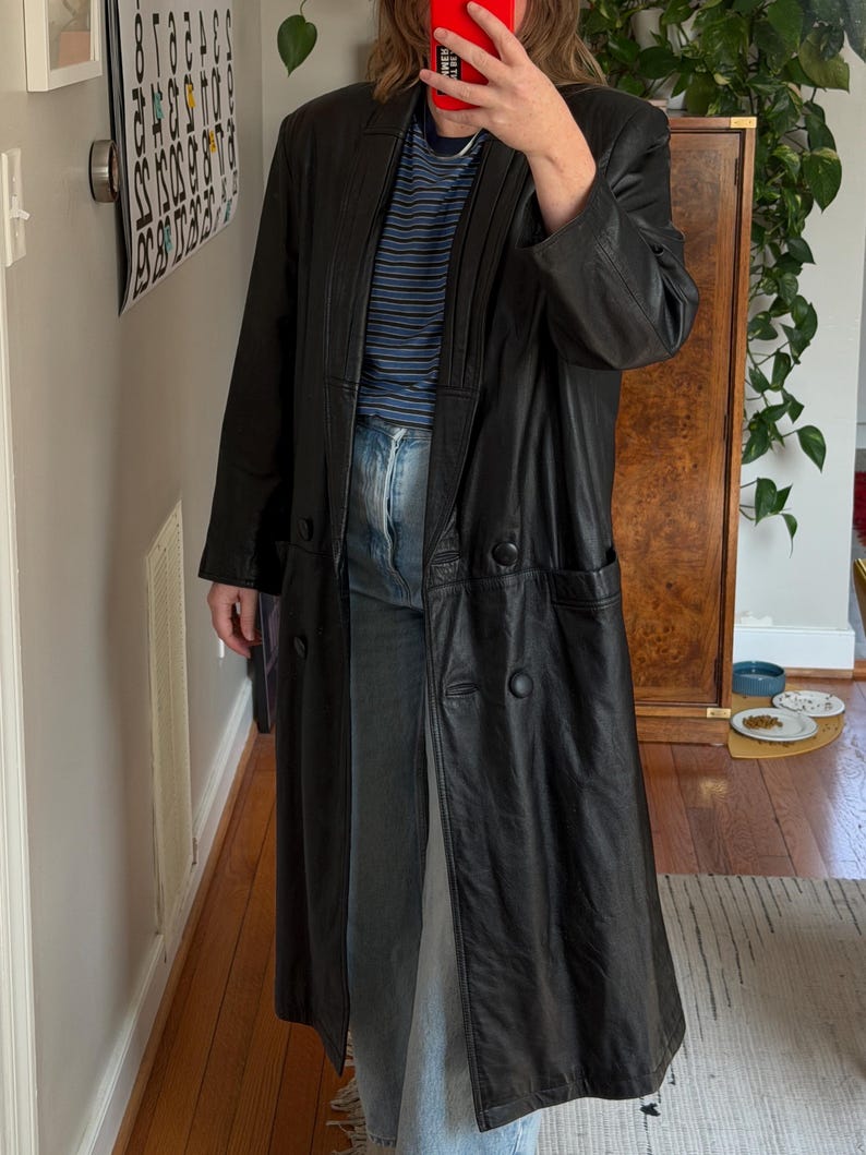 May include: A long, black leather coat with a double-breasted design. The coat has a notched collar, two front pockets, and a long, flowing silhouette. The coat is worn over a blue and white striped shirt and blue jeans.