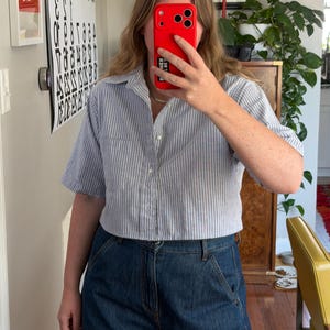 Könnte beinhalten: Ein hellblau-weiß gestreiftes Kurzarmhemd mit einer einzelnen Brusttasche. Das Hemd wird mit dunkelblauen Jeans kombiniert. Das Outfit ist lässig und für den Alltag geeignet.