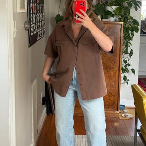 May include: A brown short-sleeved button-up shirt with two chest pockets. The shirt is paired with light blue denim jeans. The person is wearing black sandals. The outfit is casual and comfortable.