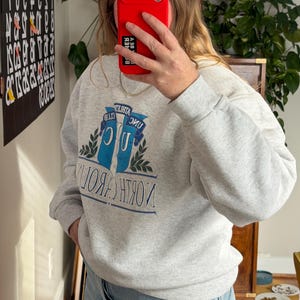May include: A light gray sweatshirt with the text "NORTH CAROLINA" in blue and white. The sweatshirt features a graphic of two blue banners with the text "ATHLETA CLUB" and a green leaf design. The sweatshirt is made of a soft material.