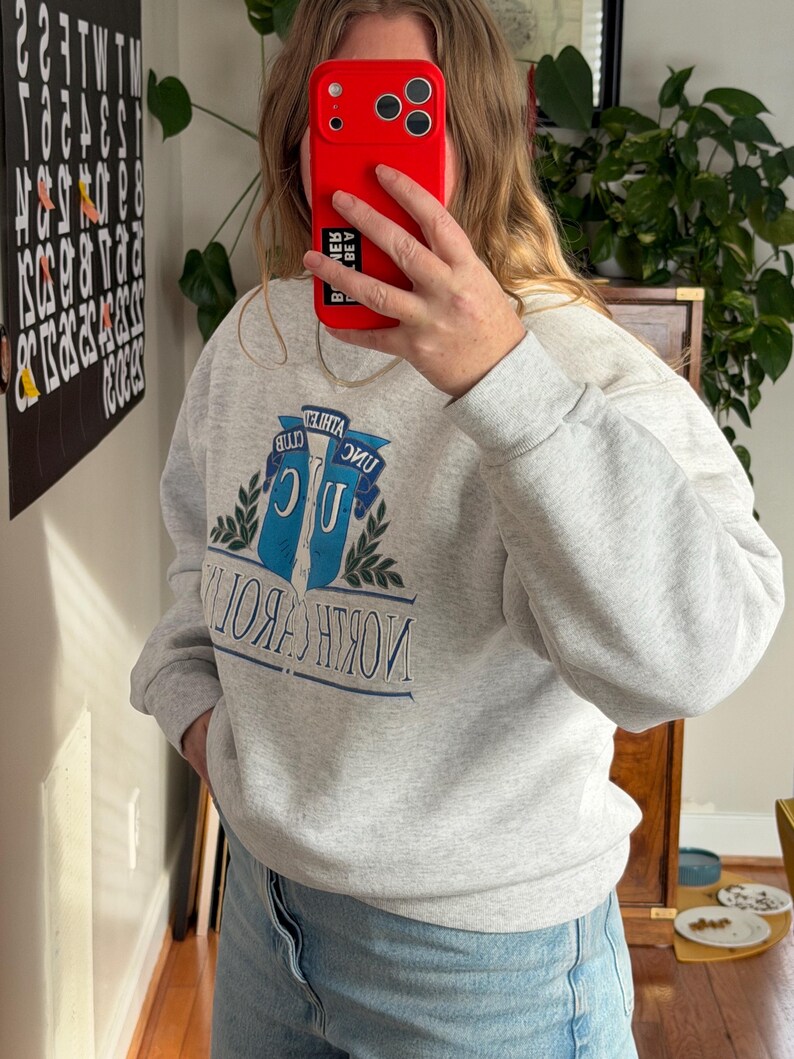 May include: A light gray sweatshirt with the text "NORTH CAROLINA" in blue and white. The sweatshirt features a graphic of two blue banners with the text "ATHLETA CLUB" and a green leaf design. The sweatshirt is made of a soft material.