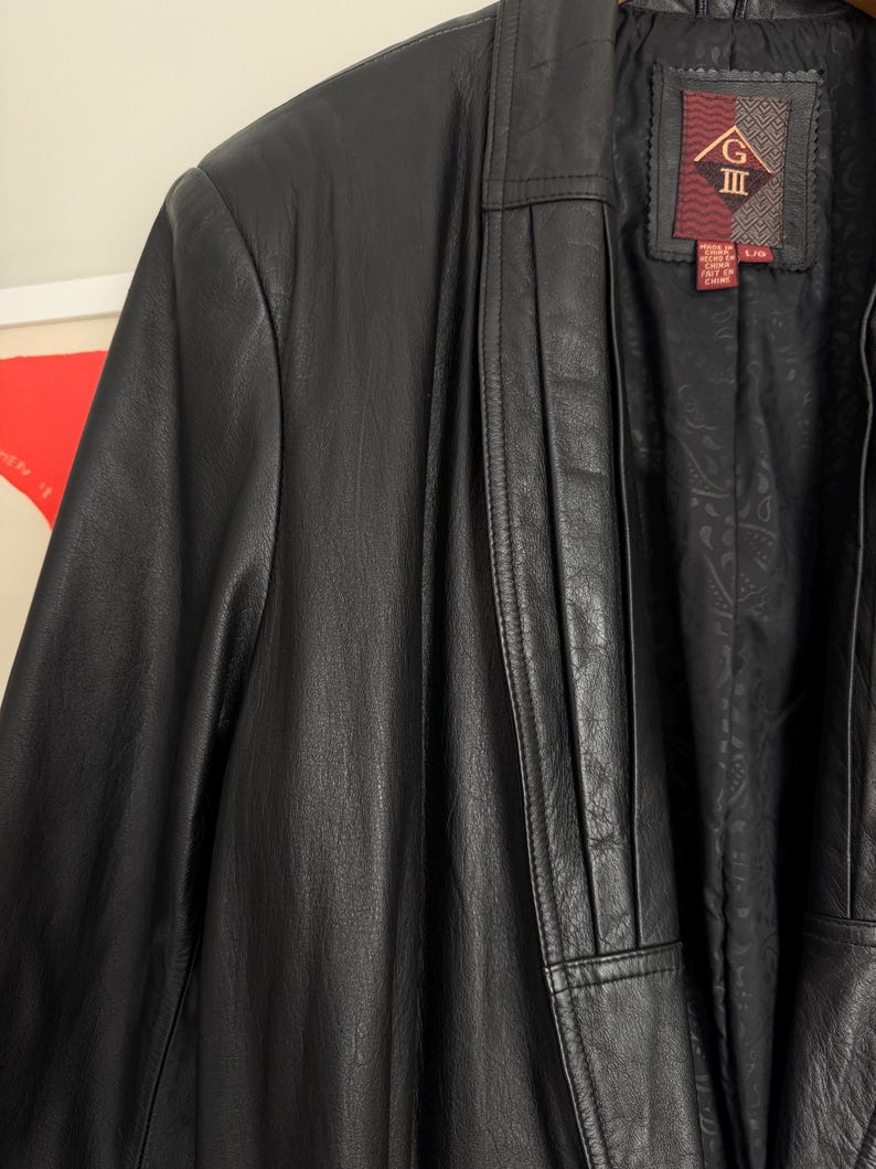 May include: Close-up of a black leather jacket. The jacket has a notched collar and a button closure. The interior lining features a paisley pattern. The label reads "G III" and includes size information.