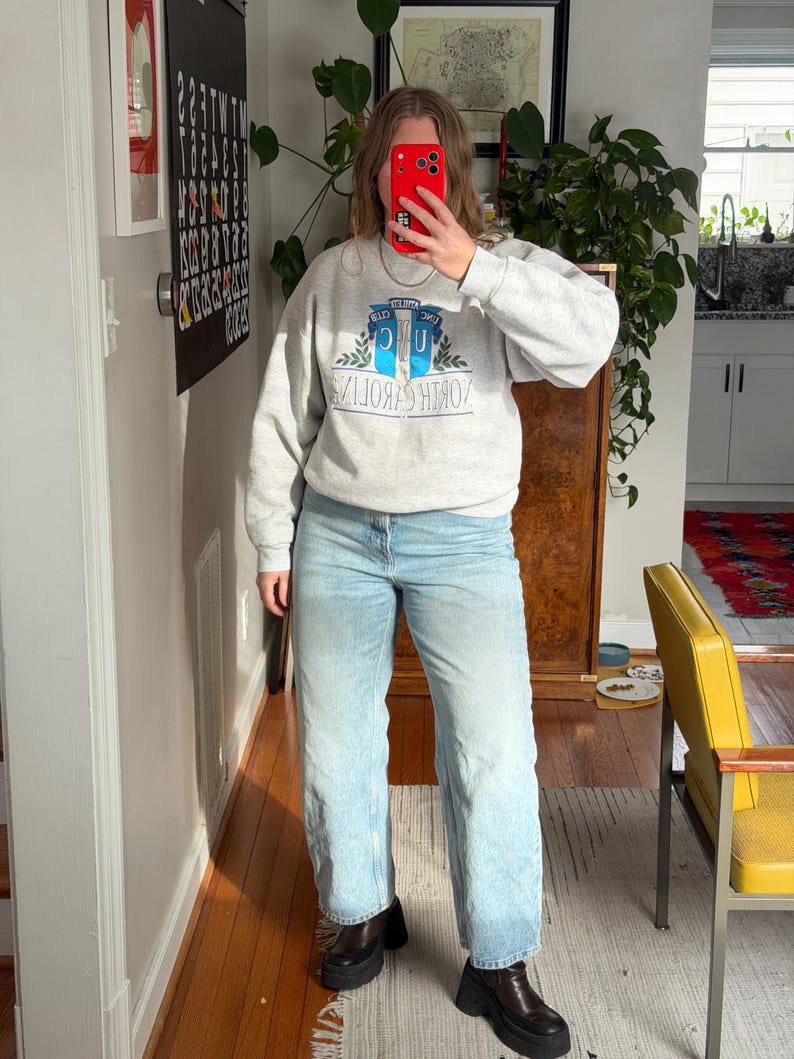May include: A light gray sweatshirt with the text "NORTH CAROLINA" in a blue and white design. The sweatshirt is paired with light blue jeans and black platform boots. The outfit is casual and comfortable.