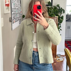 May include: A light green button-up jacket with a leaf pattern. The jacket has a collar and is worn over a white top and blue jeans. The jacket is made of a soft material.
